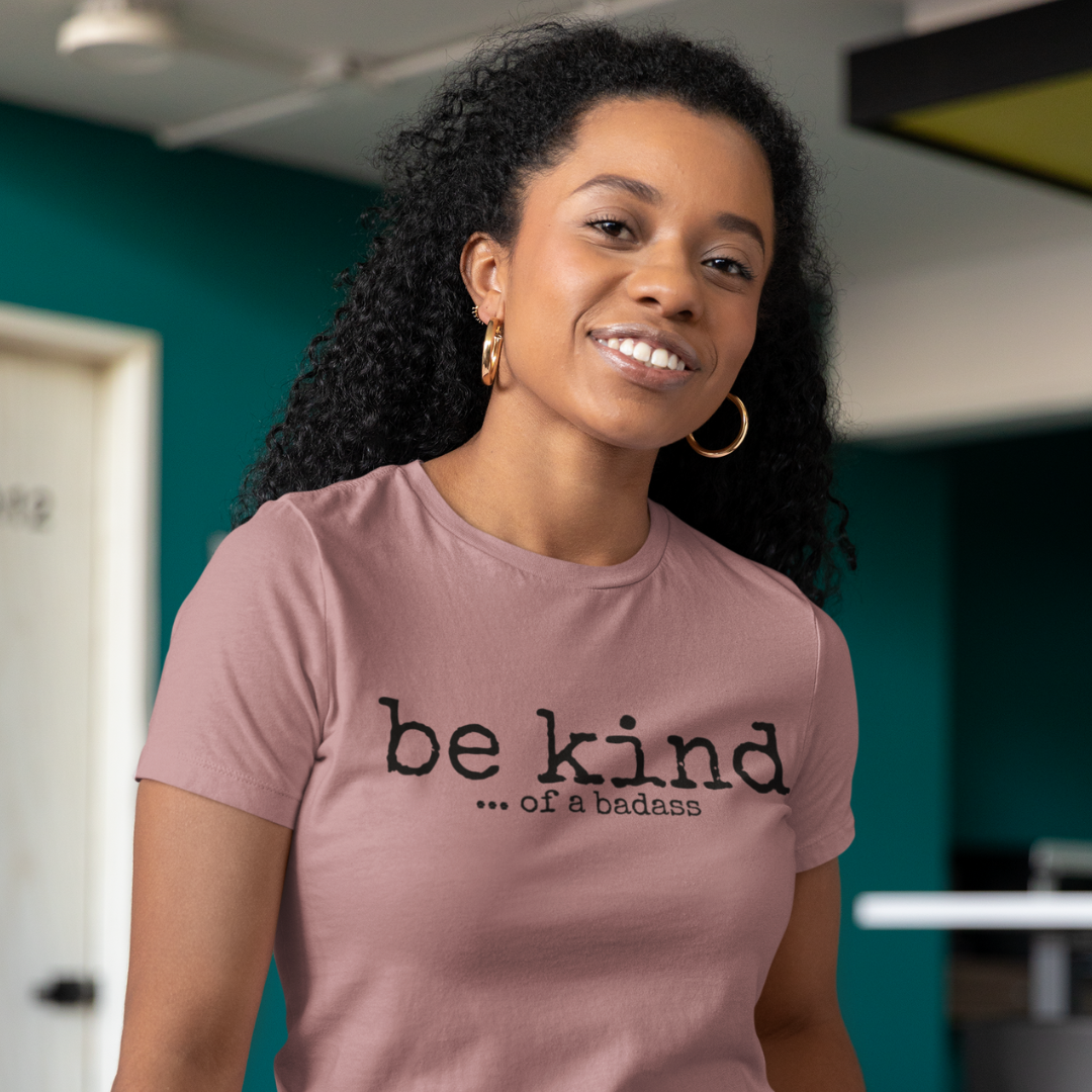 A woman radiates confidence wearing the "Be Kind ... of a Badass" T-shirt from HeadhunterGear. The playful typewriter font adds a touch of retro charm to the empowering message across the chest. 